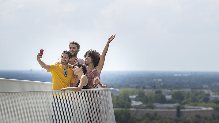 Eine Menschengruppe macht ein Selfie mit einem Handy auf dem Wolkenhain im Kienbergpark hoch über Berlin